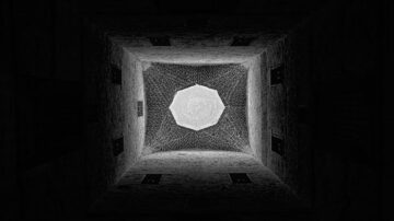 منظر علوي لزوايا مبنى مظلم يظهر فتحة مسدسة مضيئة في السقف، مع جدران حجرية مغطاة بشبكة معقدة. يبرز التباين بين الظلال والضوء.