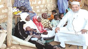 رجل مسن يرتدي ملابس تقليدية يجلس على الأرض ويتحدث مع مجموعة من النساء والأطفال في بيئة ريفية. النساء يرتدين أزياء ملونة، وبعضهن يحملن أطفالًا. الخلفية تتكون من جدران حجرية وأشخاص آخرين يجلسون بالقرب منهم، مما يعكس تفاعلًا اجتماعيًا وثقافيًا.