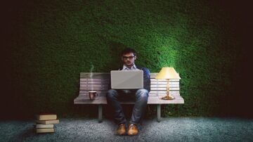 رجل يجلس على مقعد خشبي في بيئة مظلمة، يستخدم لابتوب ويعمل بتركيز. بجانبه مصباح صغير وكوب قهوة، مع كتب موضوعة على الأرض. خلفية خضراء تضفي إحساسًا بالهدوء.