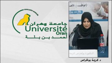 امرأة ترتدي حجاباً أسود تتحدث في مؤتمر، بجانبها زجاجة مياه. في الخلفية، شعار جامعة وهران، الجزائر.