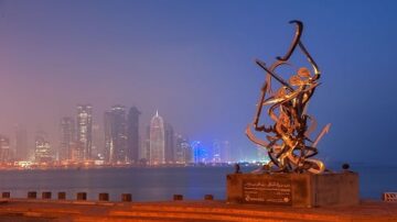 تمثال فني معاصر يظهر في الواجهة البحرية لمدينة دبي، مع أفق المدينة الحديث في الخلفية. السماء ملبدة بالغيوم بينما تتلألأ الأضواء في المباني الشاهقة. يعكس التمثال الإبداع المعماري والفني لدبي.