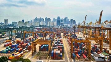 منظر جوي لميناء بحري مزدحم يحتوي على حاويات ملونة متعددة الأشكال في صفوف منظمة، مع رافعات عملاقة خلفية. تظهر skyline المدينة الحديثة في الخلفية، مما يعكس النشاط التجاري والحركة في المنطقة.