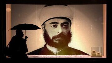 شخص يحمل مظلة يقف أمام صورة كبيرة لرجل يرتدي عمامة تقليدية، يظهر وجهه بوضوح في إضاءة خافتة. خلفية الصورة تحتوي على كؤوس بيضاء، مما يضيف لمسة فنية للمشهد.