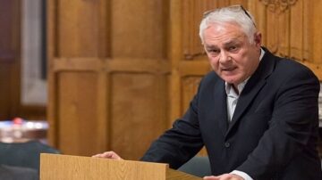 رجل مسن يرتدي سترة سوداء يتحدث من منصة خشبية في قاعة رسمية، خلفه جدران خشبية مزخرفة. تعكس تعابيره الجدية والتركيز أثناء إلقاء كلمته.