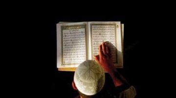شاب يقرأ القرآن الكريم في مكان مظلم، يظهر الكتاب مفتوحًا أمامه. يركز على النص بينما يلمس الصفحات بإصبعه.
