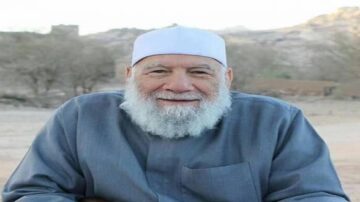 رجل مسن ذو لحية بيضاء يرتدي عمامة وملابس تقليدية، يجلس في منطقة صحراوية مع خلفية جبلية. تعبيره يدل على السعادة والود.