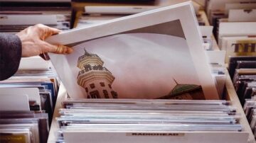 يد تظهر وهي تلتقط صورة من مجموعة من الصور الفوتوغرافية المصفوفة في صندوق. تحتوي الصور على مناظر معمارية، بما في ذلك برج مميز.