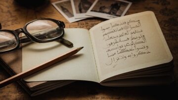 نظارة قراءة على دفتر مفتوح يحتوي على نص عربي، مع قلم رصاص بجانبه، وصور قديمة في الخلفية، تعكس جوًا من التأمل والإبداع.