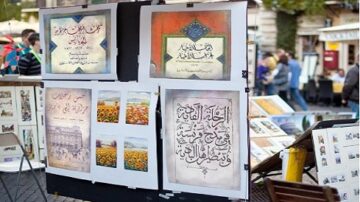 لوحة تعرض مجموعة من الأعمال الفنية، تتضمن نصوصًا خطية عربية وصورًا ملونة، مثل لوحات زهور. تعرض الأعمال في سوق أو معرض فني، مع أشخاص في الخلفية.