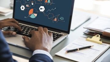 شخص يستخدم حاسوبًا محمولًا يعرض رسومات بيانية وعناصر تحليل بيانات، مع التركيز على الإحصائيات والرموز الأمنية. على الطاولة، يوجد مستندات وقلم، مما يشير إلى بيئة عمل مهنية.