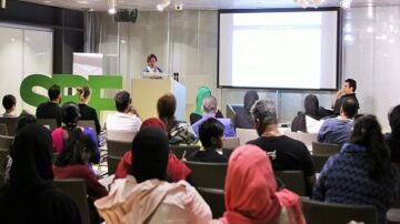 صورة لحدث تعليمي حيث يتحدث متحدث أمام جمهور متنوع في قاعة مؤتمرات. يظهر في الخلفية شعار "SPE"، وتظهر النساء والرجال في مقاعدهم بينما يركزون على العرض التقديمي على الشاشة.