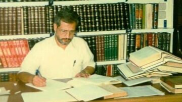 رجل يجلس على مكتب يكتب بيد ويراجع مستندات محاطة برفوف مليئة بالكتب والمجلدات. الأوراق متراكمة على الطاولة، مما يشير إلى بيئة عمل أكاديمية أو بحثية.