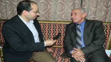 رجلان يجلسان في غرفة مزينة بأثاث تقليدي، أحدهما يحمل ميكروفونًا ويتحدث بجدية بينما الآخر يستمع بانتباه. تعكس الأجواء حوارًا مثيرًا يتناول موضوعات ثقافية أو اجتماعية.