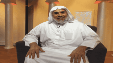 رجل مسن يرتدي الثوب التقليدي السعودي والشماغ، يجلس على أريكة في استوديو مضاء. يحتوي المكان على أعمدة مزخرفة وخلفية بسيطة، ويبدو مبتسمًا ومريحًا.