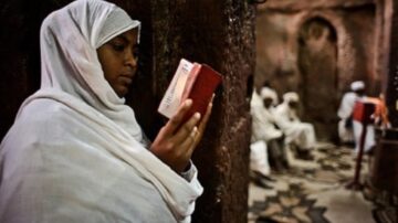 امرأة ترتدي حجابًا أبيض تقرأ كتابًا مقدسًا في كنيسة قديمة، بينما تظهر خلفها مجموعة من الأشخاص يرتدون ملابس تقليدية. الأجواء تعكس روحانية المكان والتقاليد الدينية.