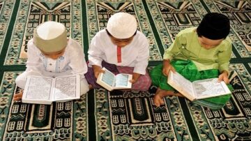 أطفال يرتدون الملابس التقليدية الإسلامية يجلسون على سجادة صلاة ويقرؤون القرآن. يظهر في الصورة ثلاثة أطفال، كل واحد منهم يحمل مصحفًا مفتوحًا، مع تفاصيل دقيقة على السجادة.