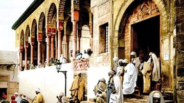 مشهد تاريخي لمجموعة من الأشخاص يرتدون أزياء تقليدية، يتجمعون أمام مبنى قديم ذو أقواس وأعمدة، مع تفاصيل معمارية مميزة. تظهر في الصورة أجواء حيوية تعكس الثقافة والتاريخ.