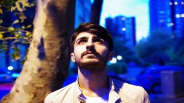 شاب ينظر إلى السماء تحت ضوء أزرق في مدينة حديثة. الأشجار والمباني تظهر في الخلفية، مما يضفي جوًا حضريًا على الصورة.