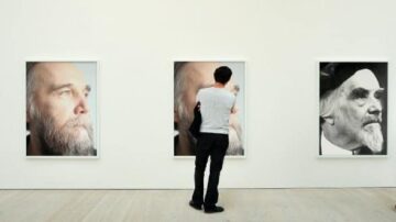 رجل يقف أمام ثلاث لوحات فنية في معرض، تعرض صوراً مقربة لوجوه رجال مسنين ذوي لحى بيضاء. تركز اللوحات على تعابير الوجه والتفاصيل الدقيقة، مع خلفية بيضاء تعزز من جمالية العمل الفني.