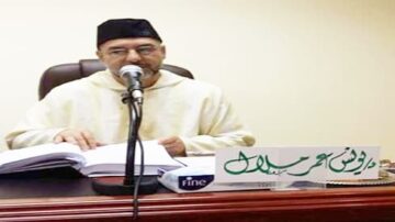 رجل يرتدي جلبابًا تقليديًا ويجلس أمام ميكروفون، يتحدث بينما يحمل كتابًا مفتوحًا. يوجد على الطاولة لافتة تحمل اسمه.