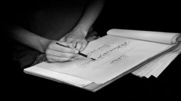 يد تظهر وهي ترسم تصميمات على ورقة بيضاء، مع دفاتر وأوراق إضافية حولها. الخلفية داكنة، مما يبرز تفاصيل الرسم.