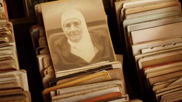 صورة قديمة لامرأة مسنّة ترتدي غطاء رأس تقليدي، تظهر في وسط مجموعة من الصور الفوتوغرافية القديمة. تبرز تفاصيل الصورة بشكل خفيف، مما يضيف طابعاً تاريخياً.