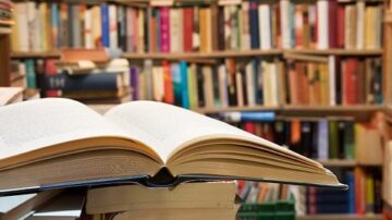 كتاب مفتوح موضوعة على طاولة، مع رفوف خلفية مليئة بالكتب الملونة. الصورة تعكس بيئة مكتبية دافئة تشجع على القراءة.