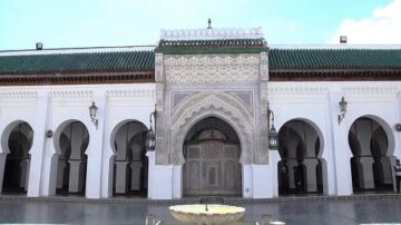 مدخل معمار تاريخي يبرز تفاصيل هندسية رائعة. يحتوي على قوس مزخرف، وأعمدة بيضاء، وسقف أخضر، مع نافورة في المقدمة. يُظهر تصميمًا مغربيًا تقليديًا يجذب الزوار.