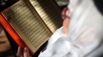 امرأة تحمل كتابًا مقدسًا، مع التركيز على الصفحات المكتوبة باللغة العربية. يظهر النص بوضوح، مما يعكس جمال الخط العربي. الضوء يسقط بشكل لطيف على الكتاب، مما يبرز تفاصيله.