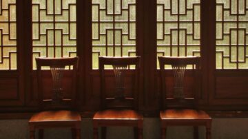 ثلاثة كراسي خشبية مصفوفة أمام نافذة ذات تصميم هندسي معقد، تسمح بدخول الضوء الطبيعي، مما يخلق جوًا دافئًا في الغرفة.