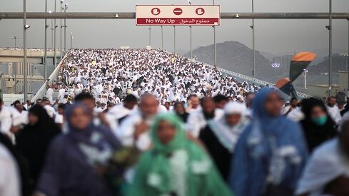 حجاج على جسر كبير في ملابس إحرام بيضاء تحت سماء غائمة وجبال في الخلف.