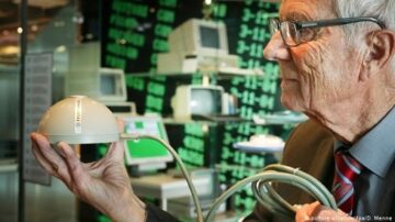 رجل مسن يحمل جهازًا دائريًا صغيرًا مع كابل في يده، بينما تظهر في الخلفية أجهزة كمبيوتر قديمة وشاشات رقمية. المشهد يعكس تطور التكنولوجيا عبر العصور.