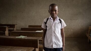 طفل صغير يرتدي زي المدرسة يقف مبتسمًا داخل فصل دراسي فارغ، مع مقاعد خشبية خلفه، وجدران رمادية عارية.