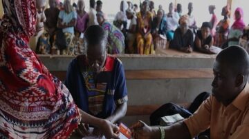 امرأة ترتدي حجابًا ملونًا تسلم بطاقة لرجل يجلس أمامها في مركز خدمات. خلفهما، مجموعة من الأشخاص يجلسون في صفوف، ينتظرون دورهم. تعكس الصورة مشهدًا من عملية تقديم الخدمات في مجتمع محلي.