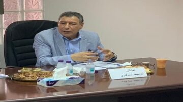 رجل مسن يجلس على طاولة اجتماعات، يرتدي سترة رمادية وقميص أزرق، وهو يتحدث ويشير بيده. على الطاولة، توجد مشروبات وعلب مناديل وحلويات. تضوء الغرفة بنور طبيعي من النوافذ خلفه.