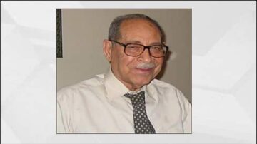 رجل مسن يرتدي نظارات وقميص أبيض مع ربطة عنق منقطة، مبتسم ويبدو وديًا، خلفية بسيطة.