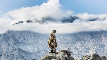 شخص يقف على صخور جبلية بملابس شتوية، خلفه جبال مغطاة بالثلوج وسحب كثيفة في السماء.