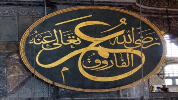 لوحة كبيرة تحمل كتابة بالخط العربي الذهبي، تتوسطها عبارة "محمد رسول الله" في مسجد آيا صوفيا. التصميم يبرز الفن الإسلامي التقليدي، مع تفاصيل معمارية مذهلة في الخلفية.