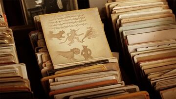 كتاب قديم يظهر رسومات لأشخاص وحيوانات، موضوعة بين مجموعة من الكتب القديمة. يبرز الكتاب في وسط الصورة بتصميمه الفريد والنقوش الموجودة عليه.