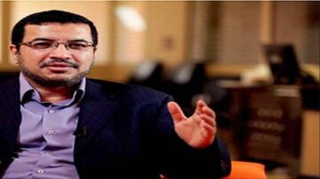 رجل في بدلة رسمية يجلس ويتحدث، مع خلفية مهنية تظهر مكاتب. يعبر عن أفكار أو آراء بأسلوب واثق، مما يعكس بيئة عمل جدية.