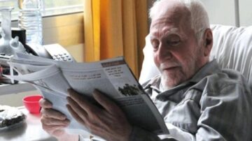 رجل مسن يجلس في سرير ويقرأ صحيفة، مع نظرة مركزة على المحتوى. الغرفة مضاءة بضوء طبيعي، وتظهر زجاجات ماء وهاتف بجواره.