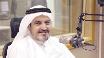 رجل يرتدي الزي التقليدي العربي يجلس في استوديو إذاعي، مبتسمًا أمام ميكروفون. خلفه زجاج يوضح بيئة الاستوديو.