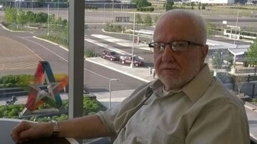 رجل مسن يجلس بالقرب من نافذة كبيرة، يرتدي نظارات وقميص بكم طويل. خلفه منظر خارجي يظهر ساحة انتظار سيارات ومجموعة من الأشجار. في الجهة اليسرى، توجد نجمة ملونة تُبرز التصميم العصري للمنطقة.