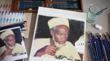 صورة لفنان يعمل على رسم صورة باستخدام أقلام تلوين، مع وجود صورة مرجعية بجانب العمل الفني. المشهد يحتوي على أدوات رسم متناثرة مثل الأقلام الملونة، والإيحاءات الفنية، وزجاجة ماء.