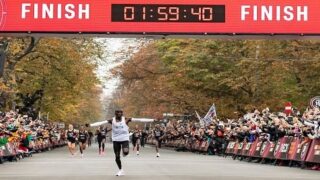 عداء يعبر خط نهاية سباق الماراثون تحت لافتة كبيرة تعرض الوقت 01:59:40، مع حشود من المشجعين على الجانبين وأشجار ملونة بالخريف في الخلفية.
