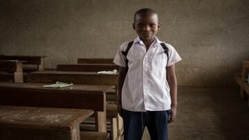 طفل يرتدي زي المدرسة يقف مبتسمًا في فصل دراسي فارغ، مع طاولات خشبية في الخلفية وجدران رمادية. يعكس الصورة بيئة تعليمية بسيطة.