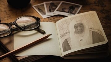 نظرة مقربة على دفتر ملاحظات مفتوح يحمل صورة شخصية لرجل مسن يرتدي عمامة، مع نظارات وقلم رصاص موضوعين بجانبه. خلفية الصورة تظهر مجموعة من الصور الفوتوغرافية القديمة.