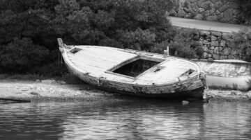 زورق قديم مهجور على ضفاف الماء، محاط بالنباتات الخضراء. يظهر في الصورة تفاصيل الخشب المتآكل والصدأ، مما يعكس حالة الإهمال.