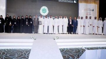 مجموعة من الأشخاص يرتدون الزي التقليدي العربي يقفون على خشبة المسرح خلال حدث رسمي. يظهر في الصورة رجال ونساء، مع وجود شعار في الخلفية. الأجواء تشير إلى مناسبة احتفالية أو تكريم.