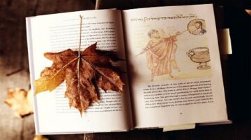 كتاب مفتوح على طاولة خشبية، مع ورقة شجر جافة موضوعة فوقه. الصفحة تعرض نصوصًا قديمة ورسومات توضيحية. تبرز الألوان الدافئة للخريف، مع أوراق شجر ملونة حول الكتاب.